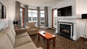 A living room with a fireplace, a sofa, a chair, a television, and large windows with curtains. - Sommet des Neiges (Mont-Tremblant)
