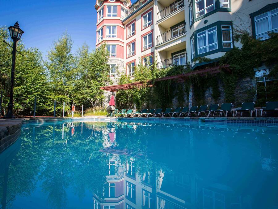 A clear swimming pool with lounge chairs and a lamp post, surrounded by trees and buildings with balconies.