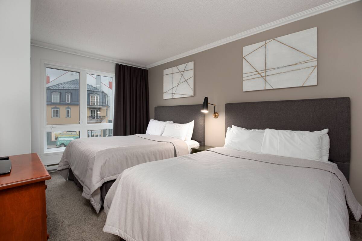 A hotel room with two beds, a desk, and a window with a view of buildings.