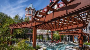 A pool area with a pergola structure, surrounded by lush greenery and a stone building in the background. - Sommet des Neiges (Mont-Tremblant)