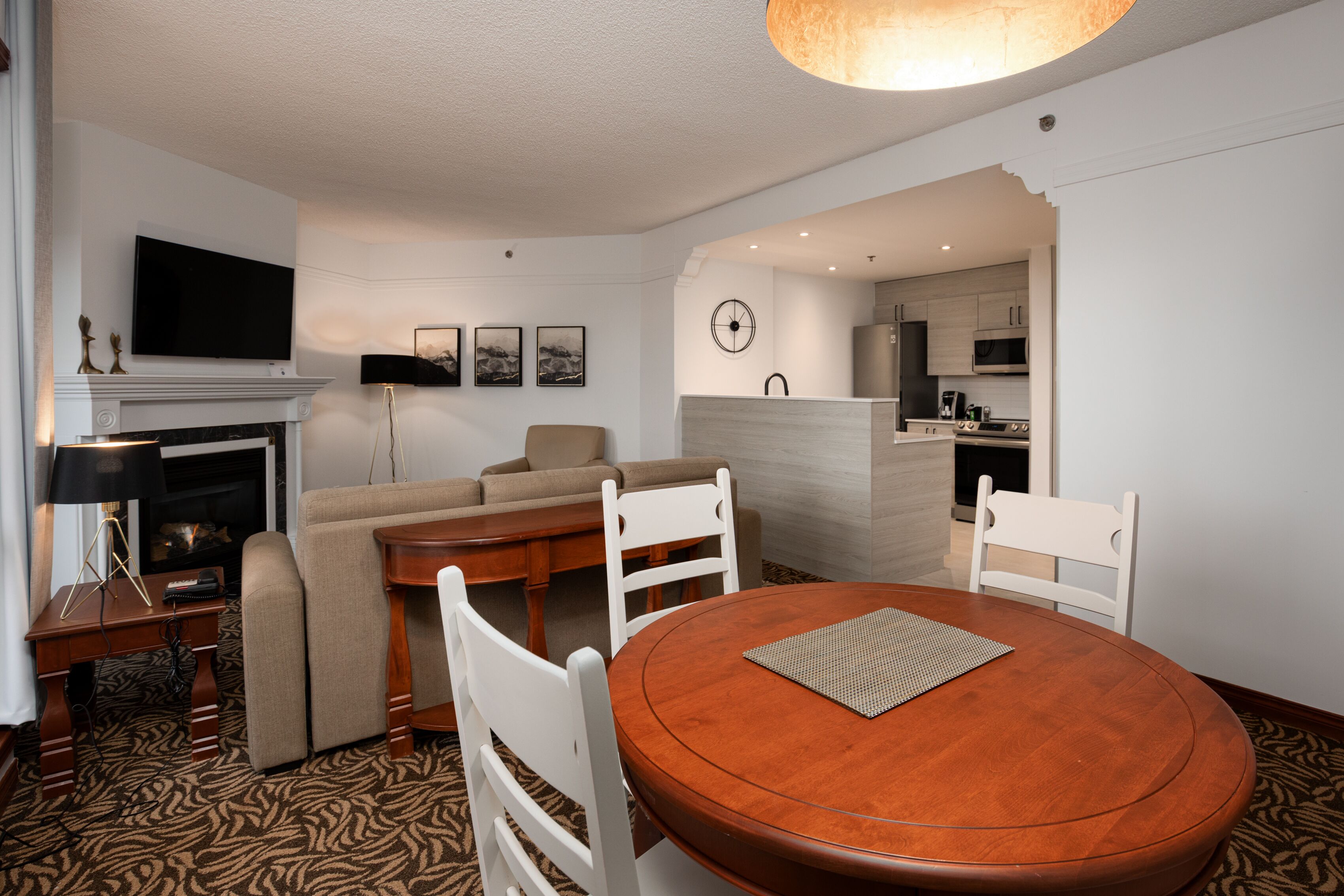 Un salon moderne avec une cheminée, un téléviseur, un coin repas avec une table et des chaises en bois, et une cuisine en arrière-plan.