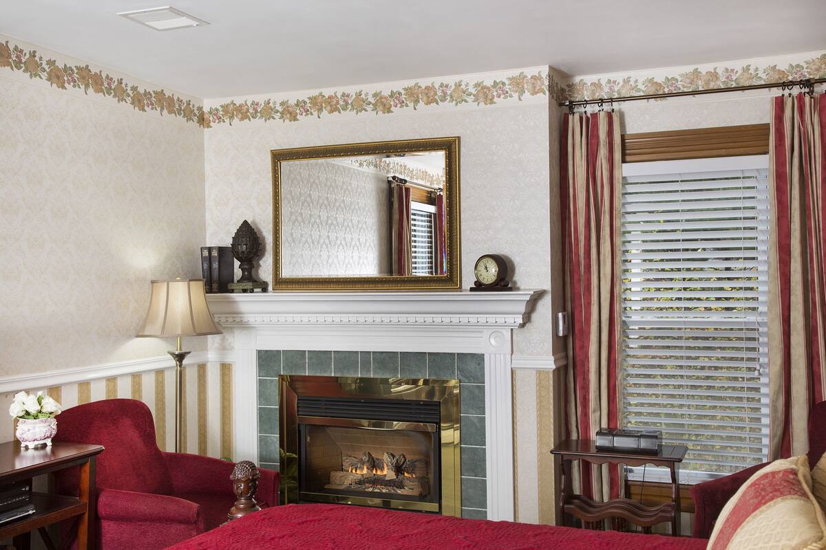 Una habitación de hotel con chimenea, un espejo grande, cortinas a rayas y una cama con una colcha roja.