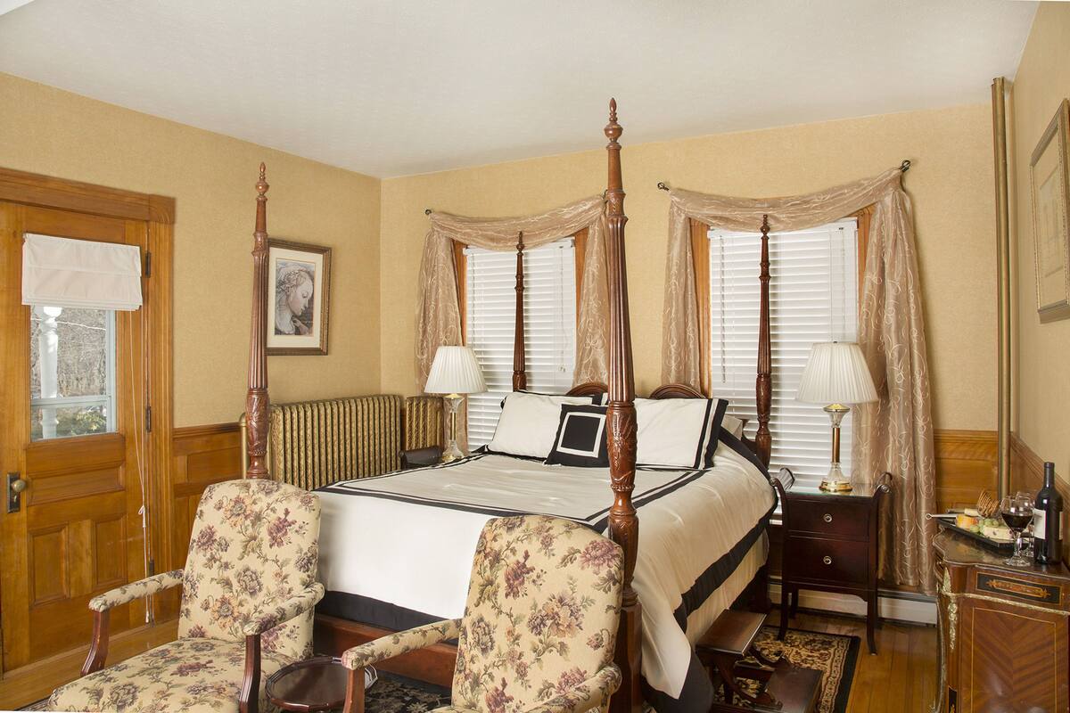 Una cama con dosel de madera, dos sillones tapizados con estampado floral, una mesita de noche con lámpara y un aparador con espejo.