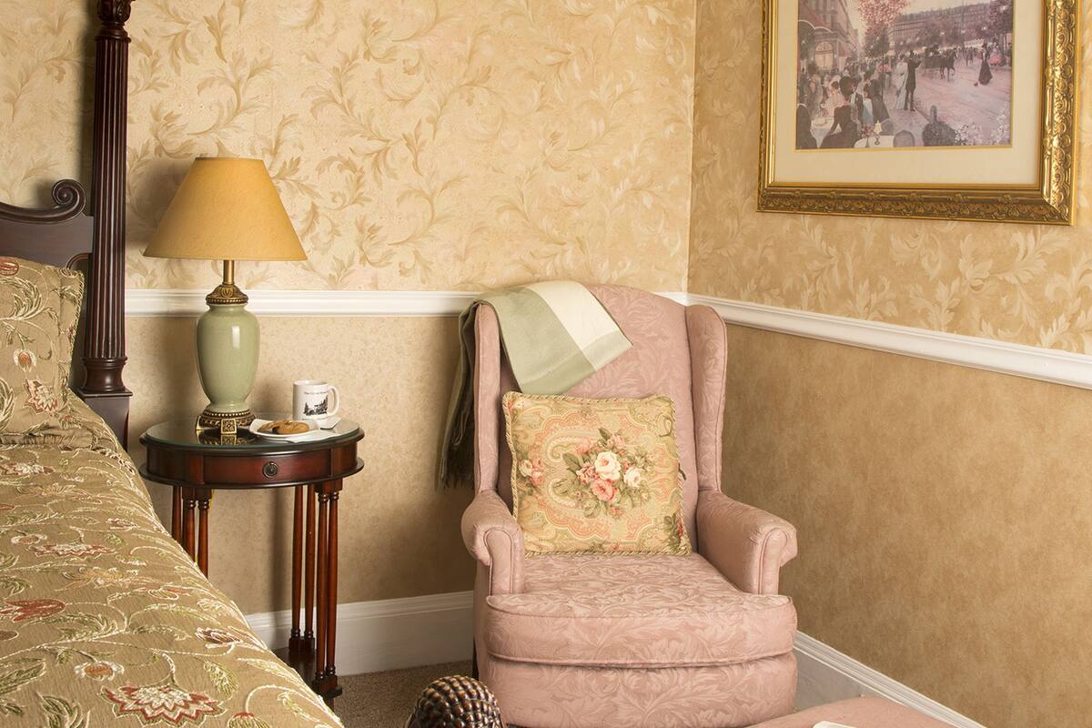 Una habitación con una cama, una silla, una mesita con lámpara y un cuadro en la pared.