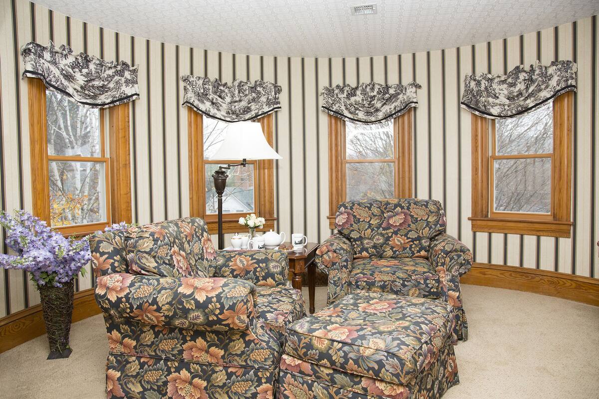 Una habitación con papel tapiz a rayas, sillones de estampado floral, una mesa con lámpara y un jarrón de flores.