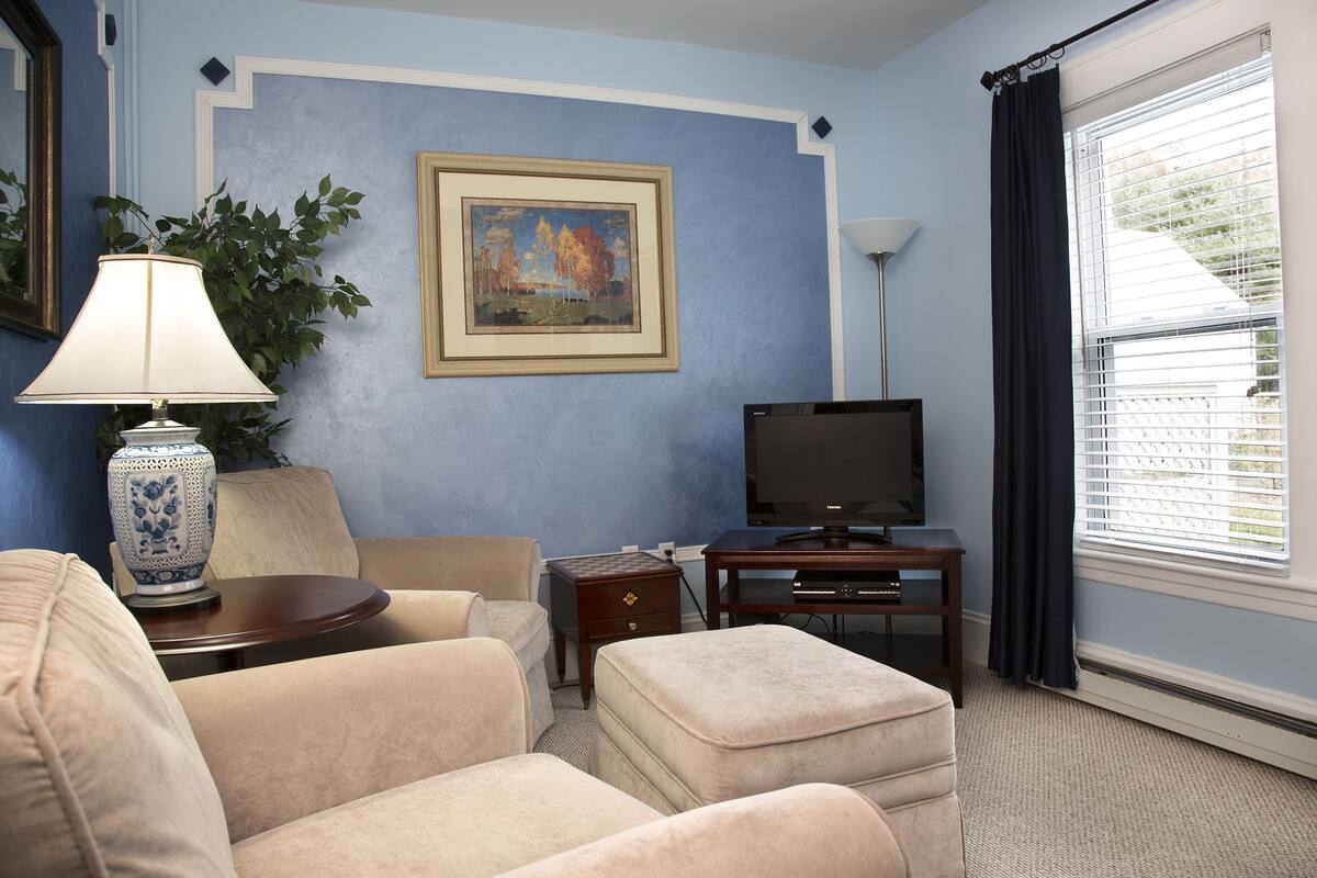 Una sala de estar con un sofá color beige, una mesita con una lámpara, un televisor y un cuadro en la pared.