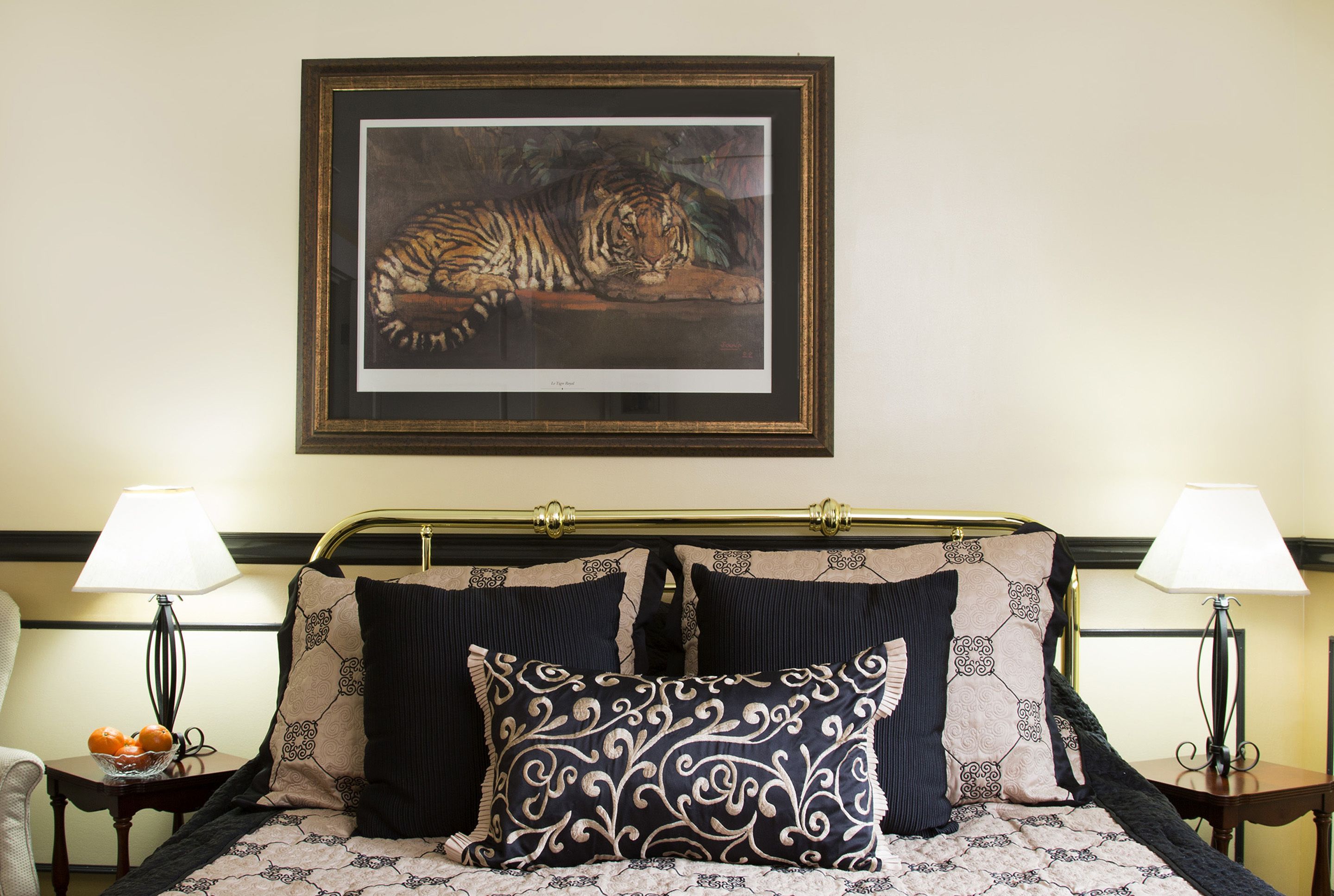 A neatly made bed with patterned pillows, a framed tiger painting above it, and two bedside lamps on either side.