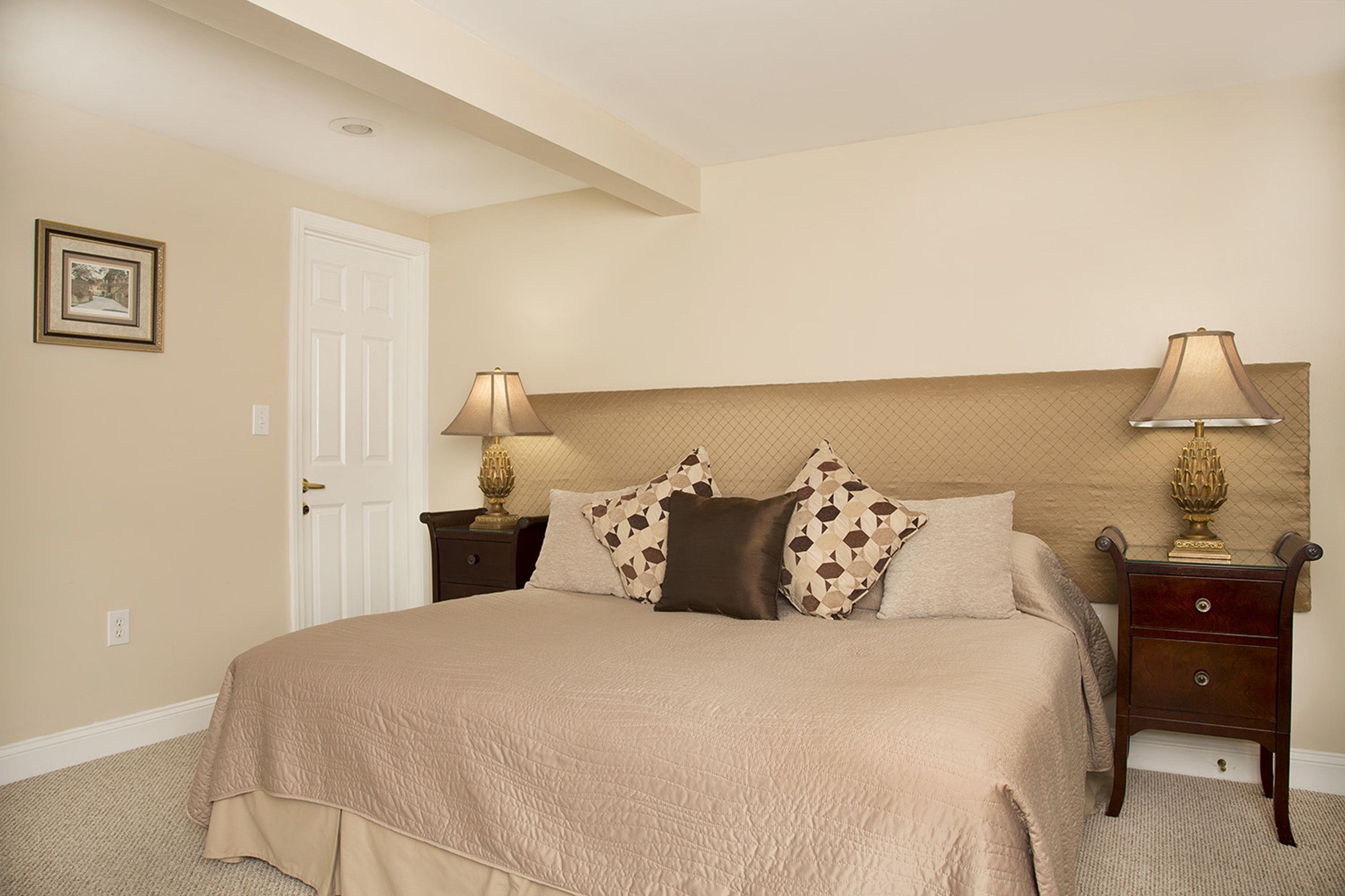 A bedroom with a large bed, two bedside tables with lamps, a door, and a framed picture on the wall.