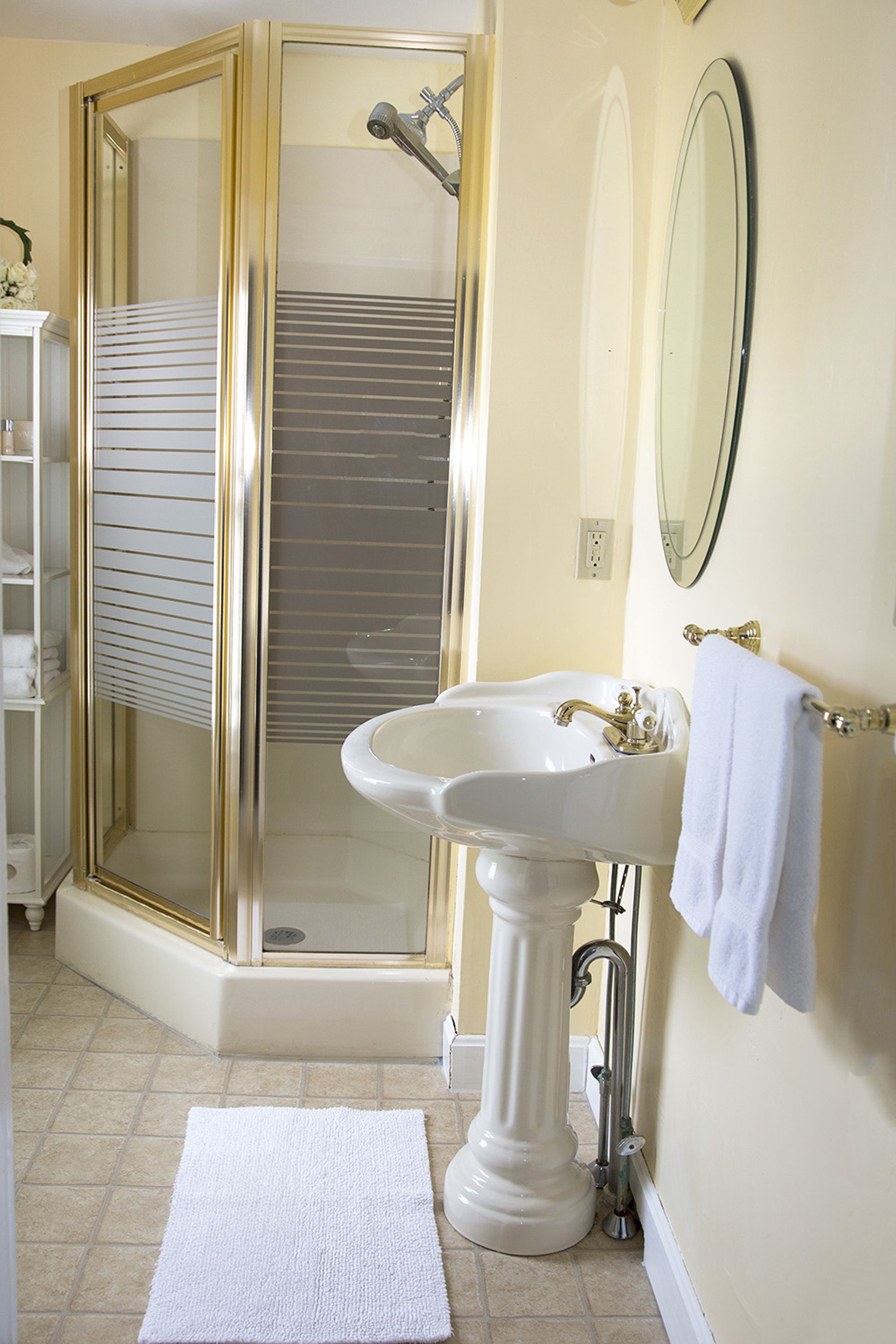 Un baño con ducha envuelta en vidrio, lavamanos pedestal, espejo y una toalla blanca en la pared.