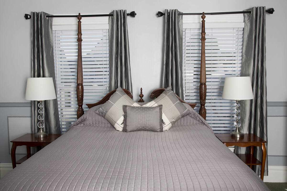 Una cama bien hecha con un edredón gris, dos mesitas de noche con lámparas, y un ventanal con persianas y cortinas.