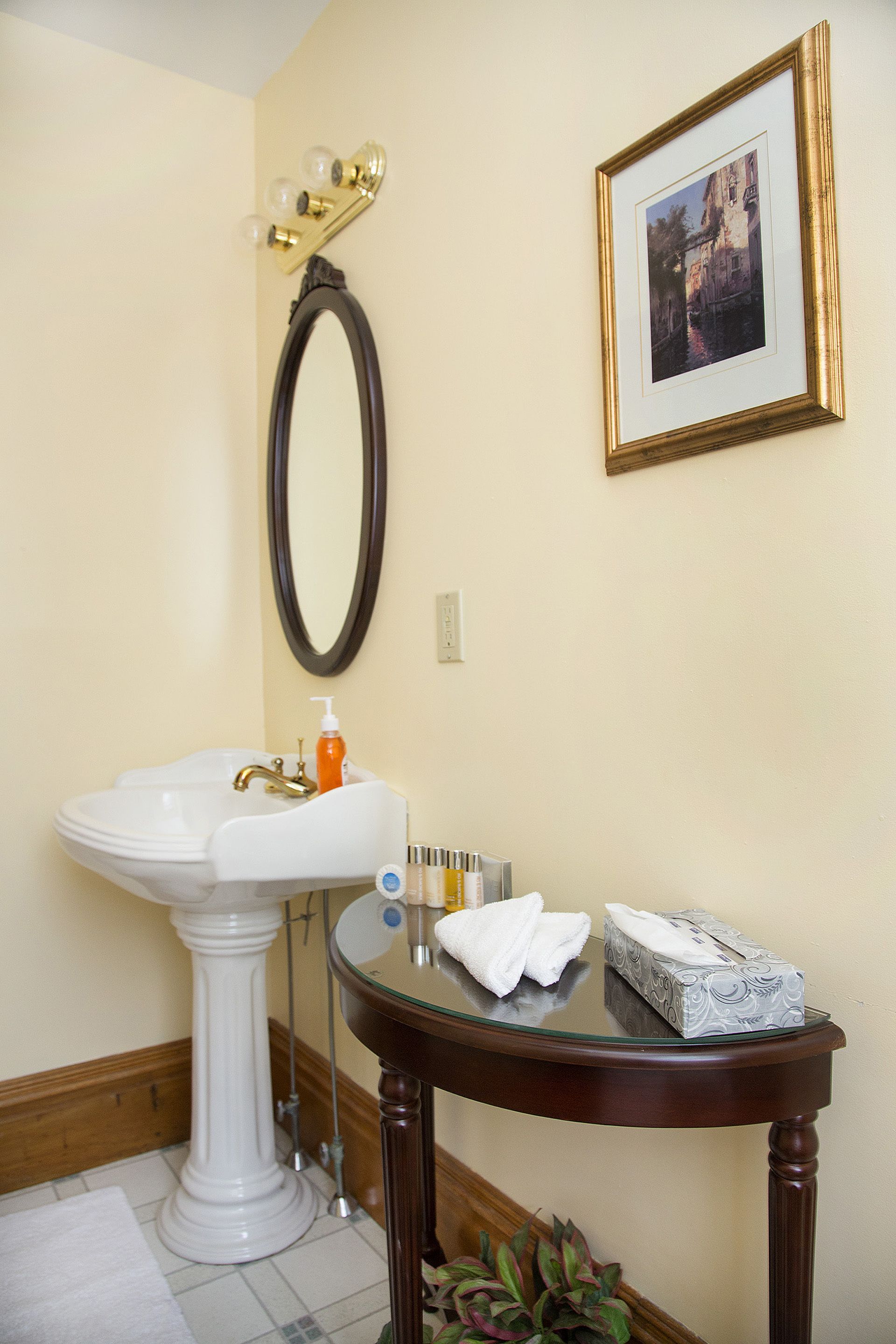 A bathroom with a pedestal sink, a round table with toiletries, a framed picture on the wall, and a mirror above the sink.