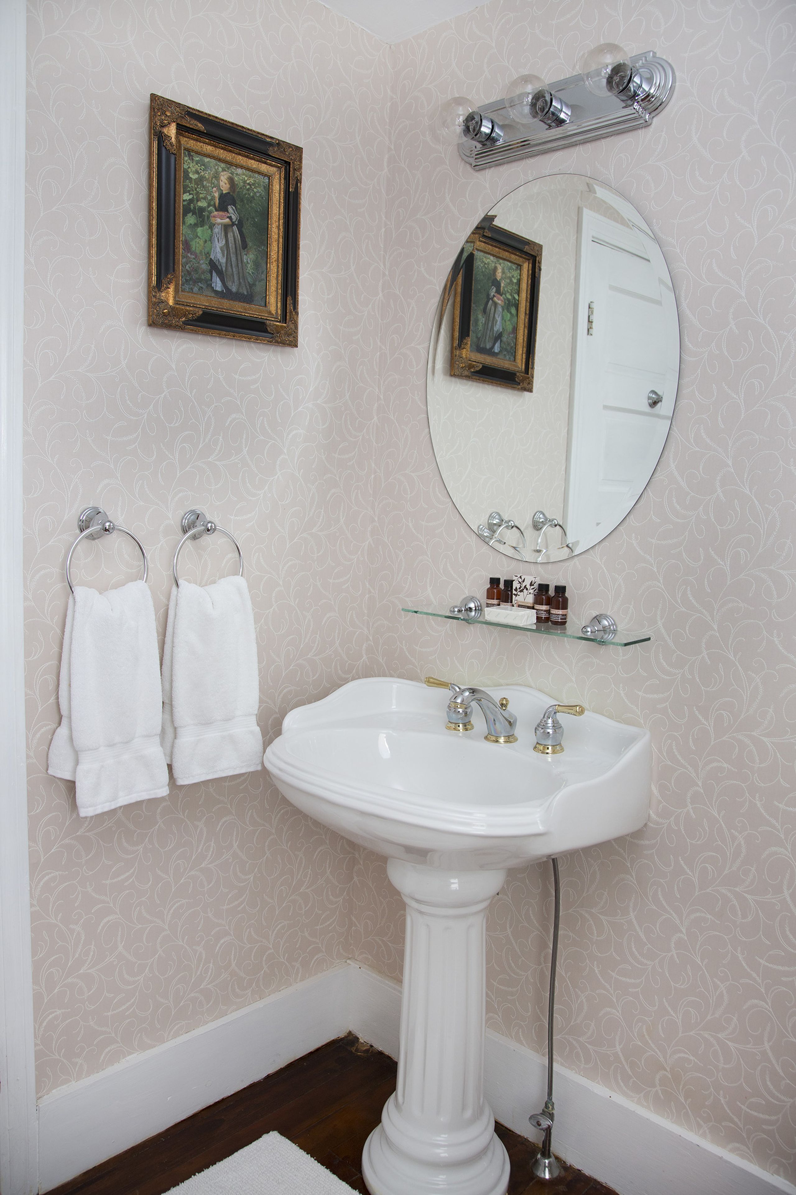 A bathroom with a pedestal sink, two wall-mounted towels, a framed picture, a mirror, and a glass shelf with toiletries.