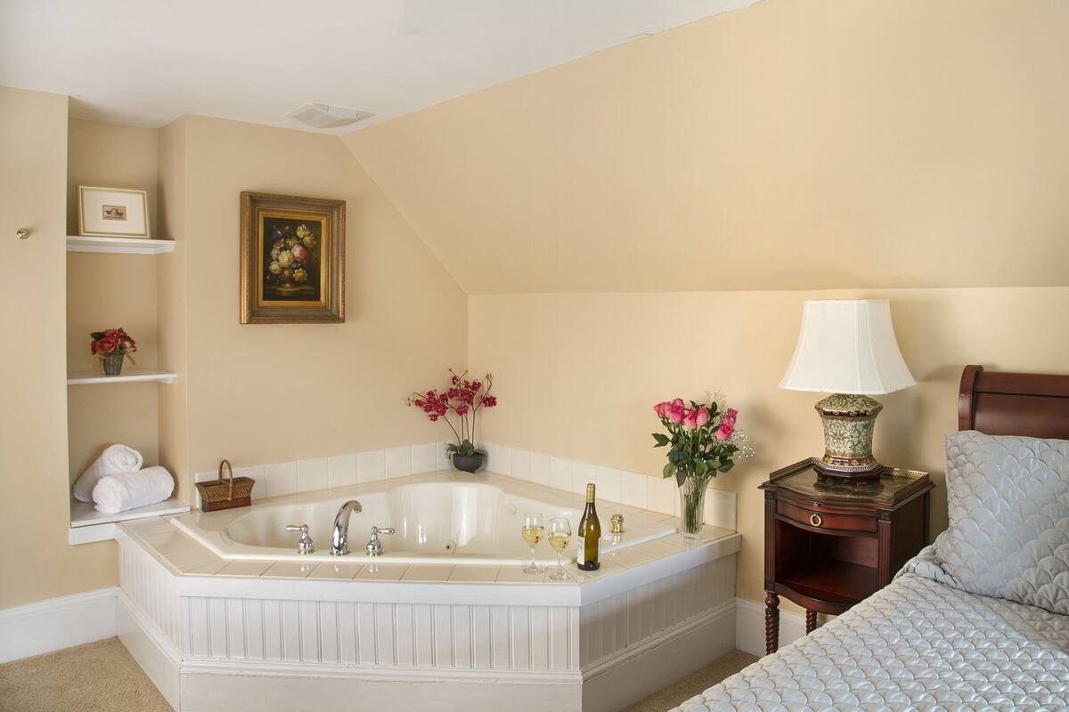 Un dormitorio con una bañera grande, una mesita de noche con lámpara y una cama con colcha acolchada.