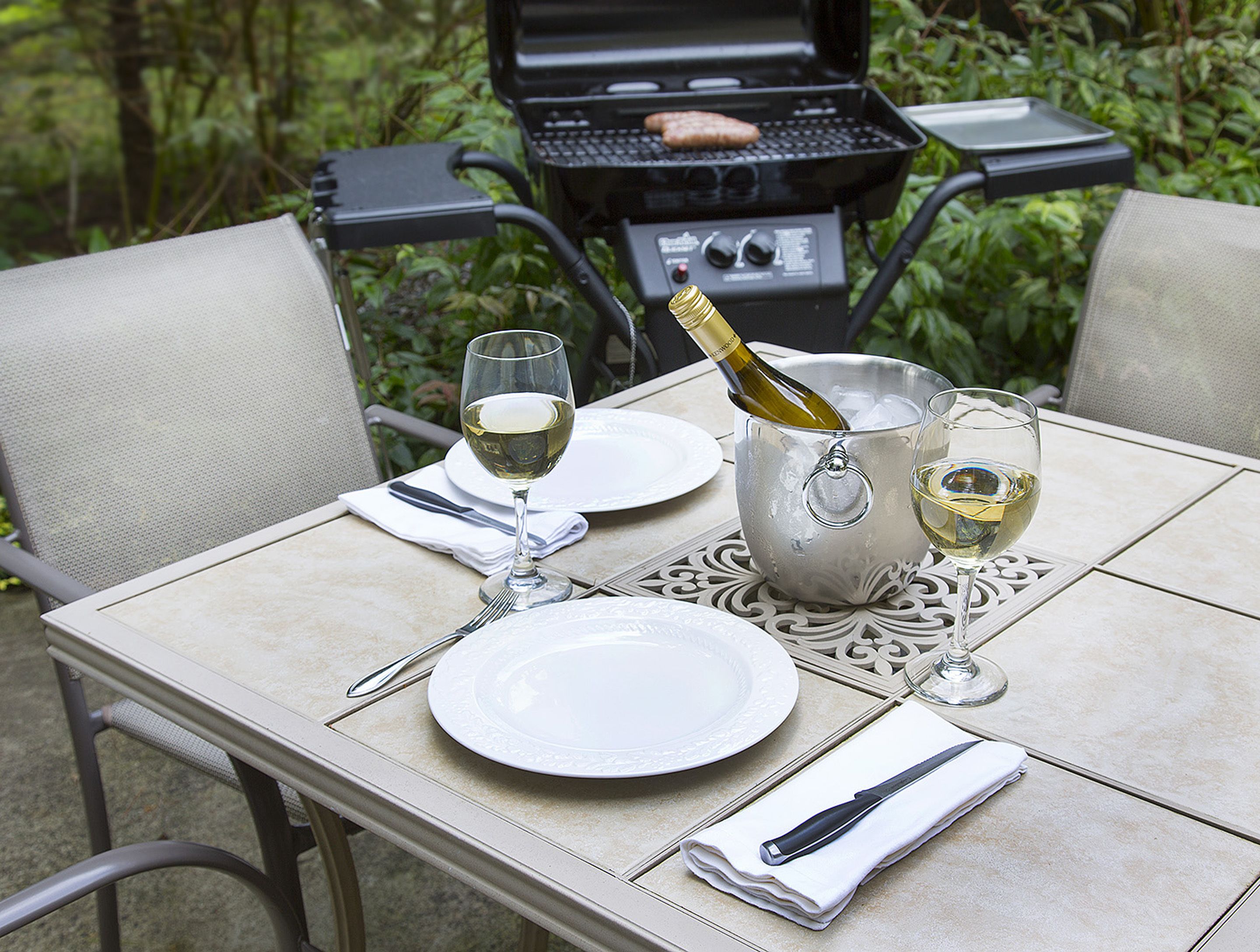 A table set for two with a bottle of wine, glasses, and a grill in the background.