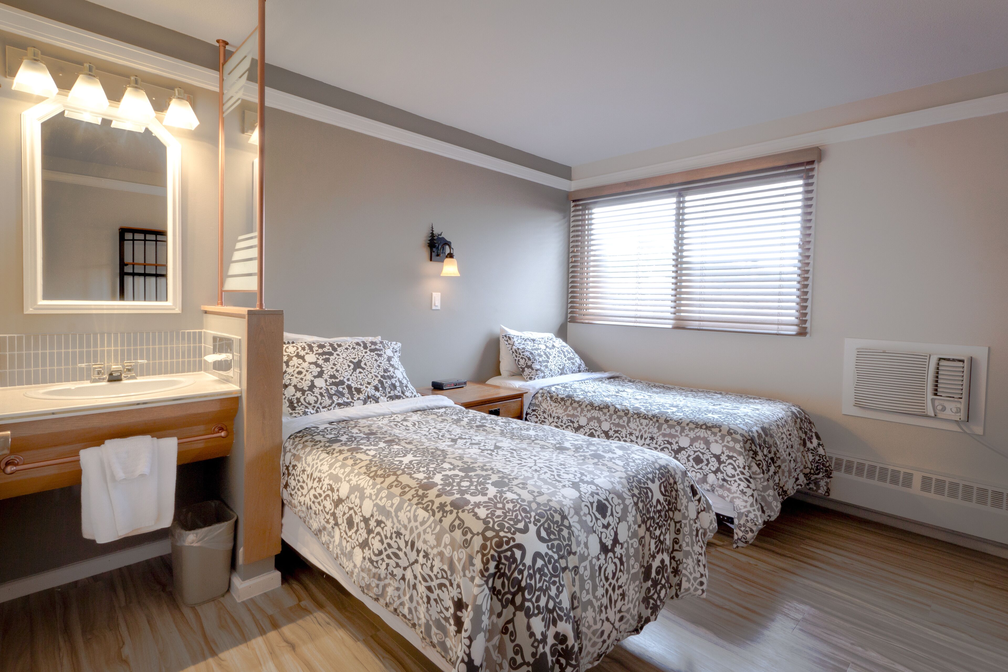 Ein Schlafzimmer mit zwei Betten, einem großen Spiegel, einem Schminktisch mit Waschbecken und einem Fenster mit Jalousien.