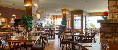 A spacious restaurant with wooden tables and chairs, a stone fireplace, and a large plant.