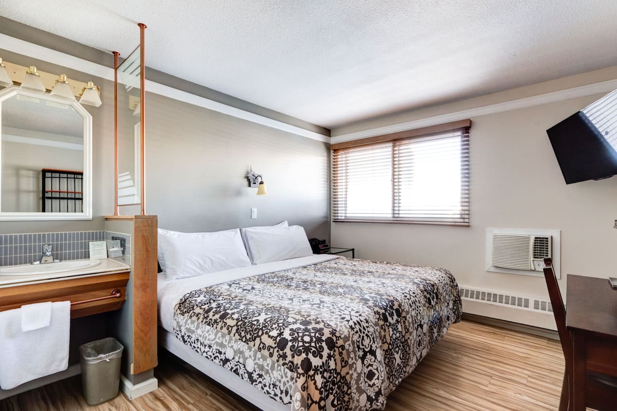 Une chambre d’hôtel avec un grand lit, une table de chevet en bois, un miroir, une fenêtre avec des stores et un climatiseur fixé au mur.