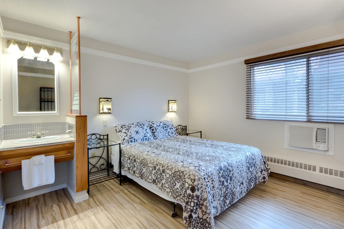 Une chambre à coucher comprenant un lit, une table de chevet, un miroir, une coiffeuse avec un lavabo et une fenêtre avec des stores.