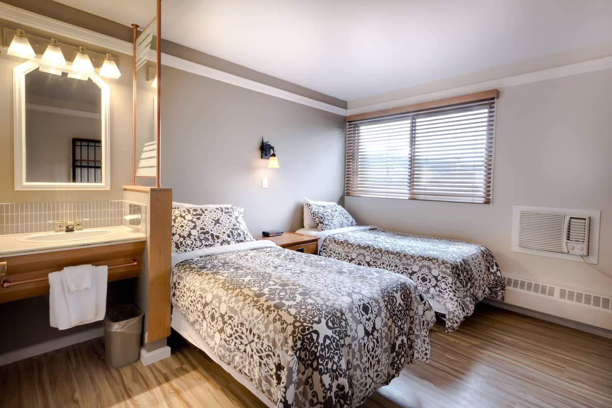 Une chambre d’hôtel avec deux lits, un grand miroir, une salle de bain avec un lavabo et une fenêtre avec des stores.