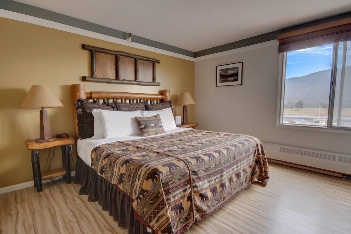 Une chambre à coucher avec un grand lit, deux tables de chevet, une tête de lit, une fenêtre donnant sur un beau paysage et un tableau accroché au mur.