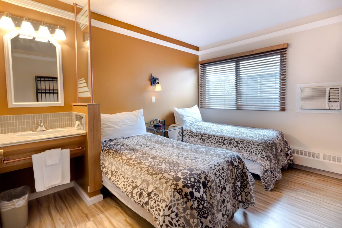 Une chambre d’hôtel avec deux lits, une salle de bain équipée d’un lavabo et d’un miroir, et une fenêtre avec des stores.
