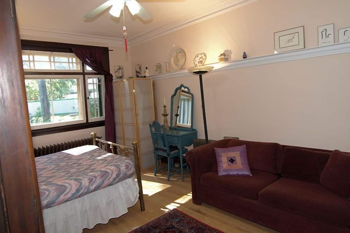 Une chambre à coucher comprenant un lit, un bureau, une chaise, un canapé rouge, un ventilateur de plafond et une fenêtre avec des rideaux.