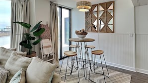 A modern living room with a sofa, a dining table with stools, a hanging light fixture, and a view of the sea through large windows. - Sunbird Beach Resort by Resort Collection (Panama City Beach)