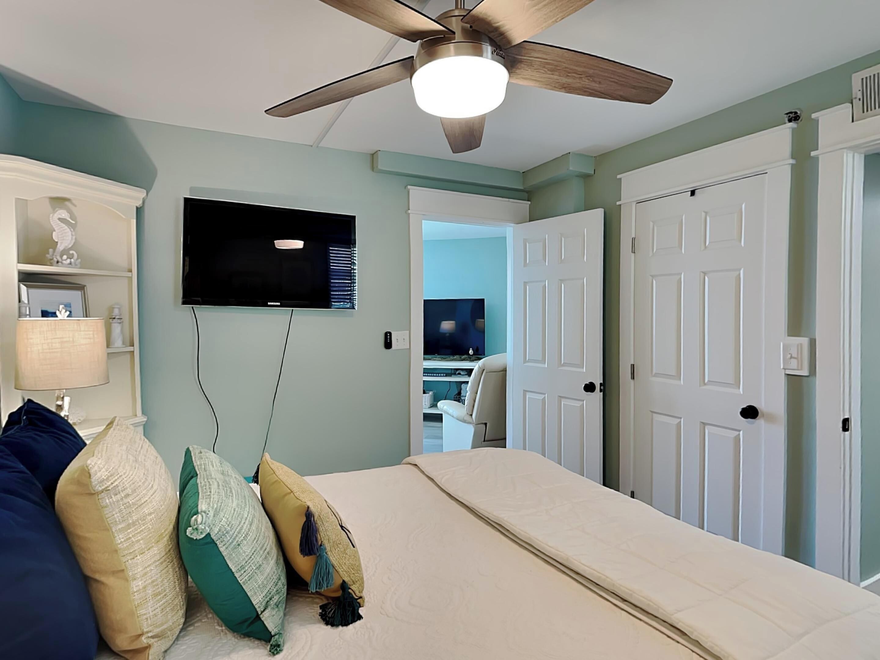 Un dormitorio con cama, ventilador de techo, televisor y puerta que da a otra habitación.