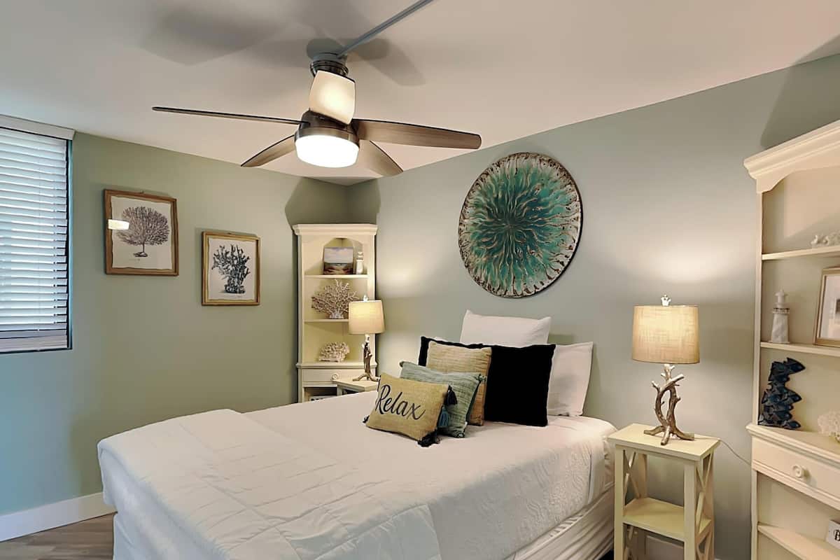 Une chambre à coucher comprenant un lit, un ventilateur de plafond, une table de chevet, une lampe et un élément décoratif mural.