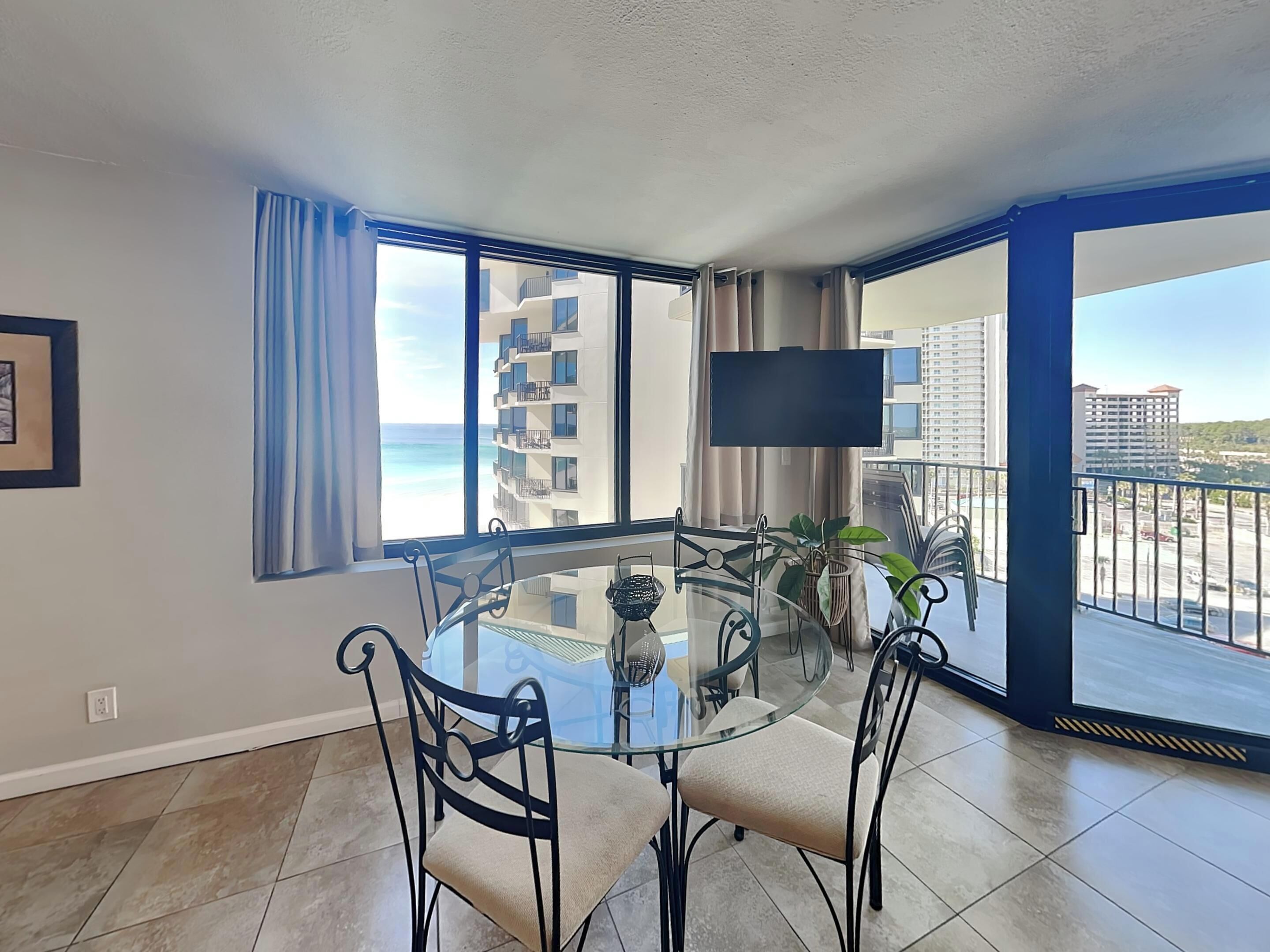 A modern dining area with a glass table and metal chairs, large windows with curtains, a flat-screen TV, and a view of the ocean and buildings.
