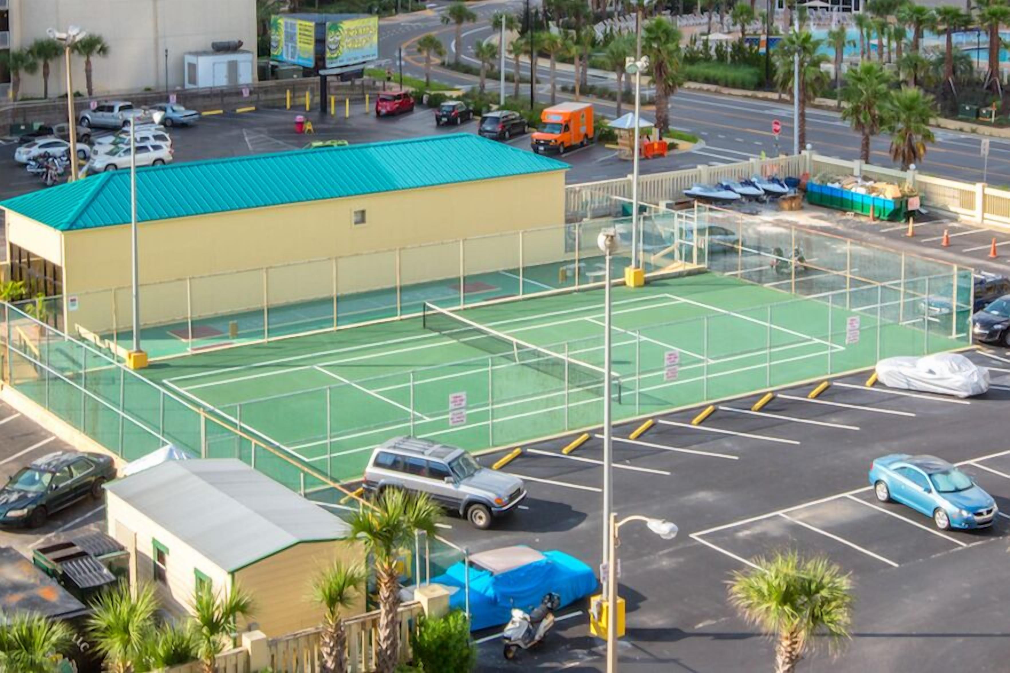 A tennis court with a green surface, surrounded by a fence, and adjacent to a building with a yellow roof.