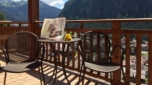Una terraza de madera con dos sillas de mimbre y una mesa, con vistas a un paisaje montañoso bajo un cielo despejado.