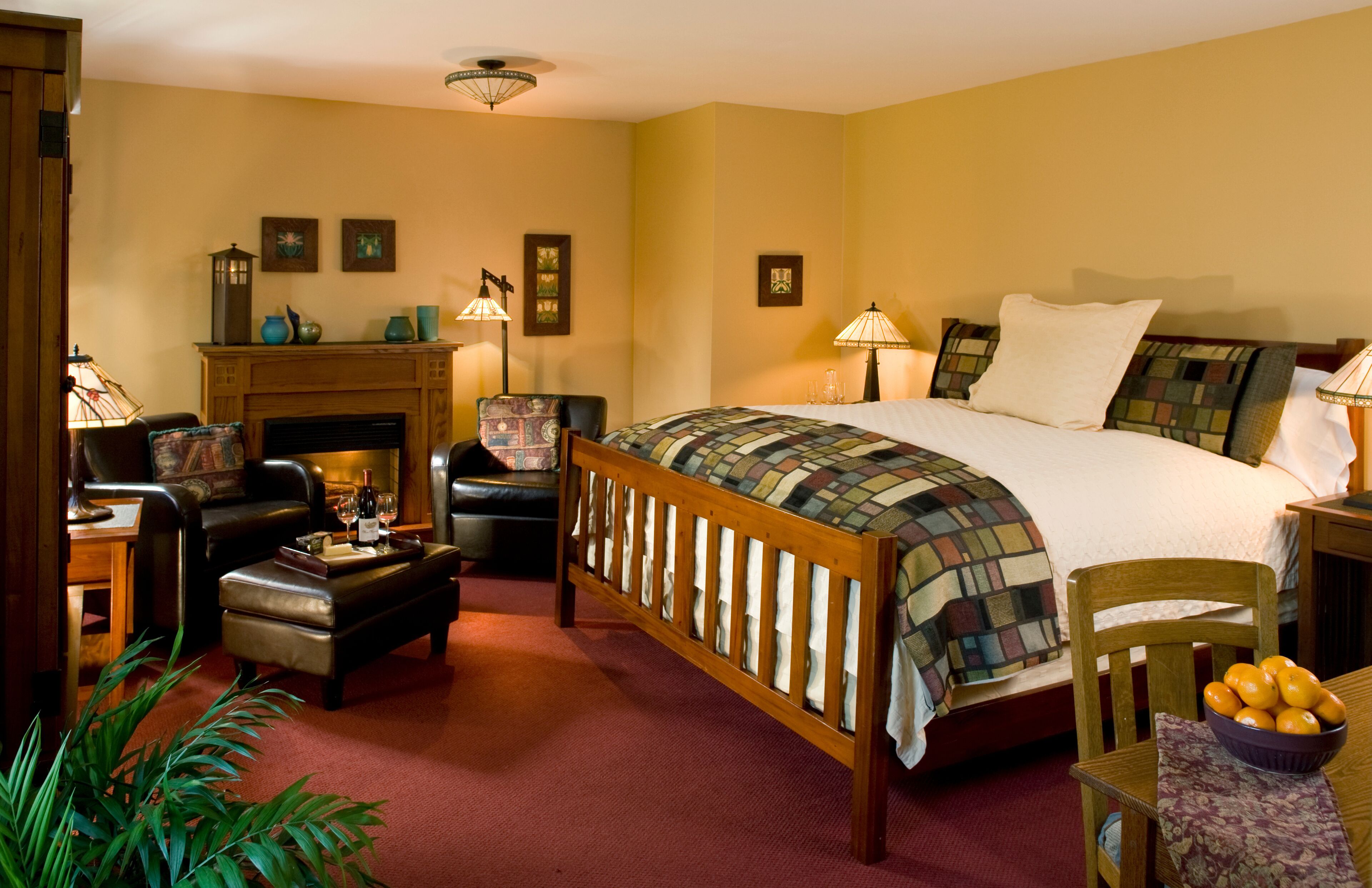 A bedroom with a large bed, a wooden chair, a small table with a bowl of fruit, a fireplace, and wall-mounted lamps.