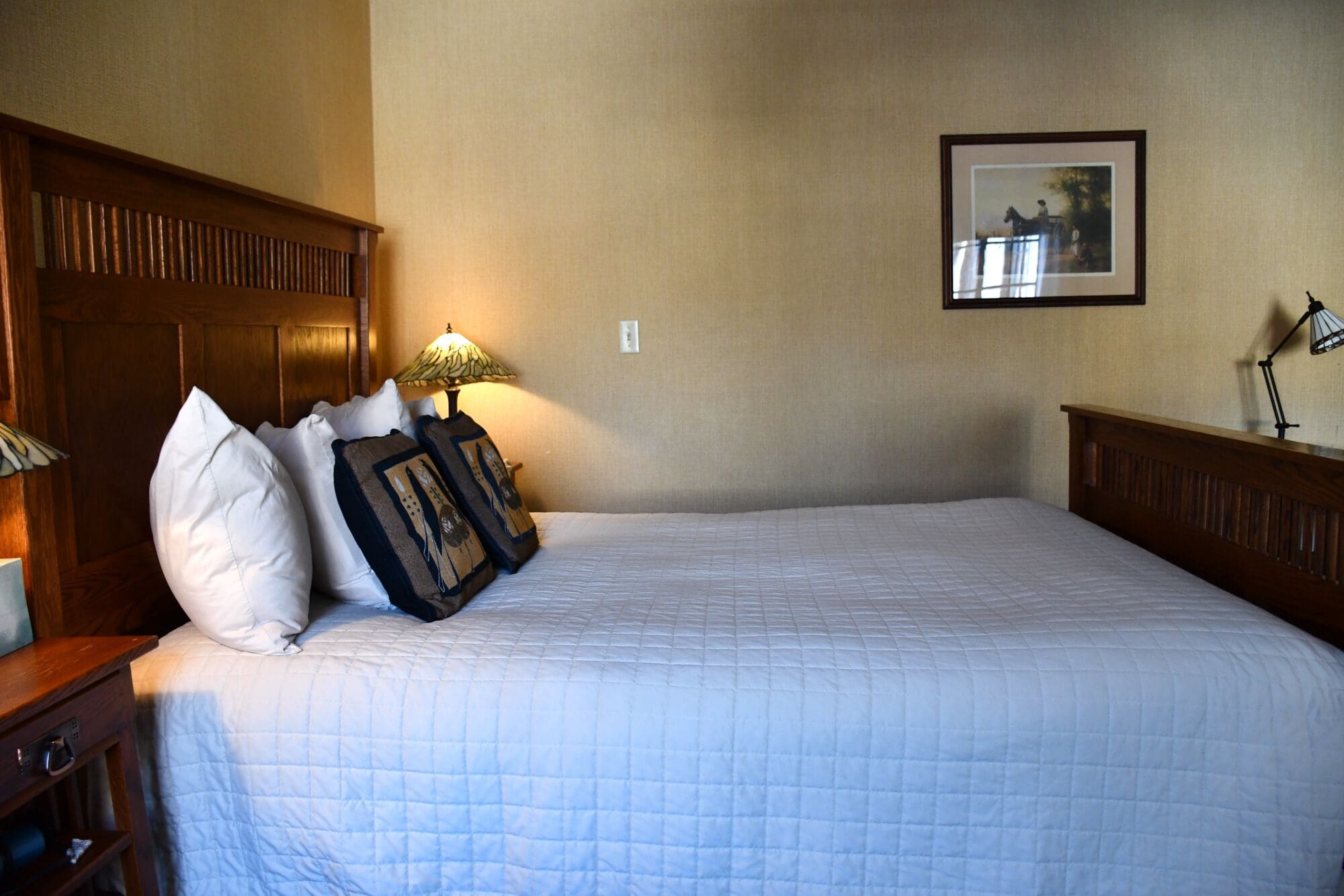 A neatly made bed with a quilted white bedspread, a wooden headboard, and two pillows with a patterned cover. A table lamp with a green shade is on the nightstand beside the bed.
