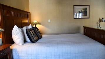 A neatly made bed with a quilted white bedspread, a wooden headboard, and two pillows with a patterned cover. A table lamp with a green shade is on the nightstand beside the bed.