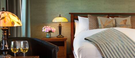 A hotel room with a bed, a nightstand with a lamp, a vase with flowers, and two glasses of wine on a table.
