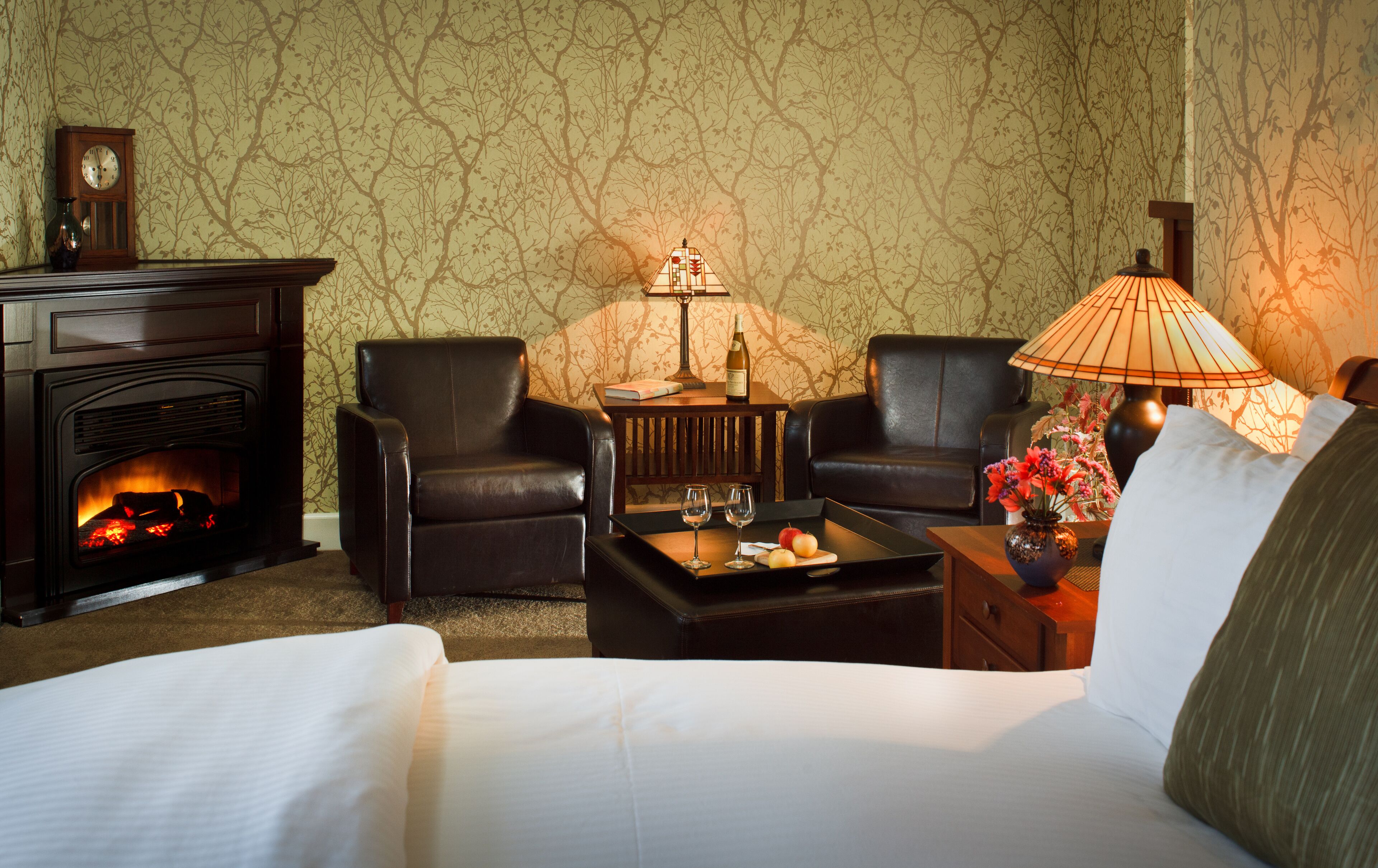 Una habitación de hotel con chimenea, dos sillones de cuero, una mesa de centro con copas de vino, una lámpara y una cama.