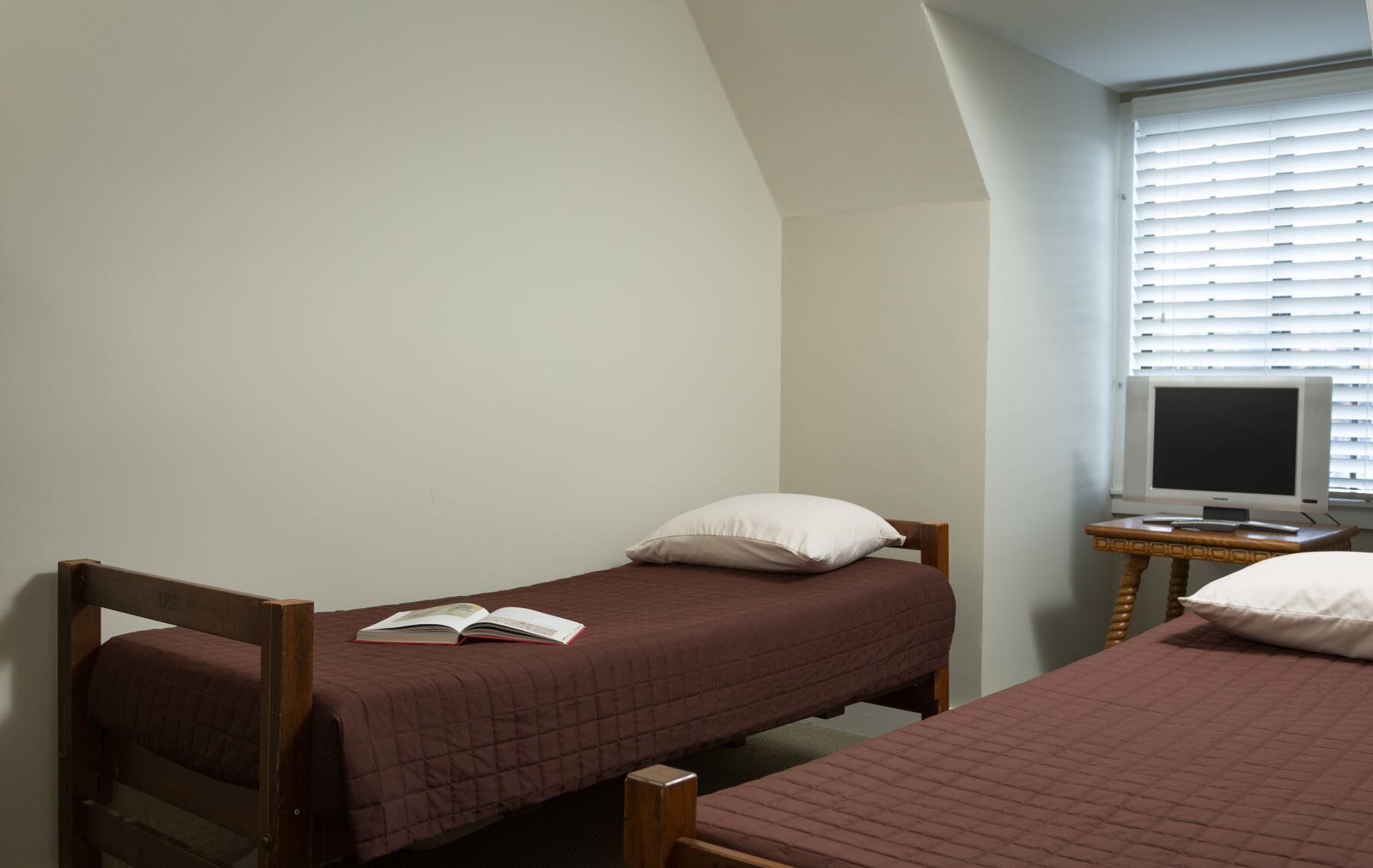 Een kleine, eenpersoons accommodatie met twee eenpersoonsbedden, een houten stoel, een klein tafeltje met een televisie en een raam met zonwering.
