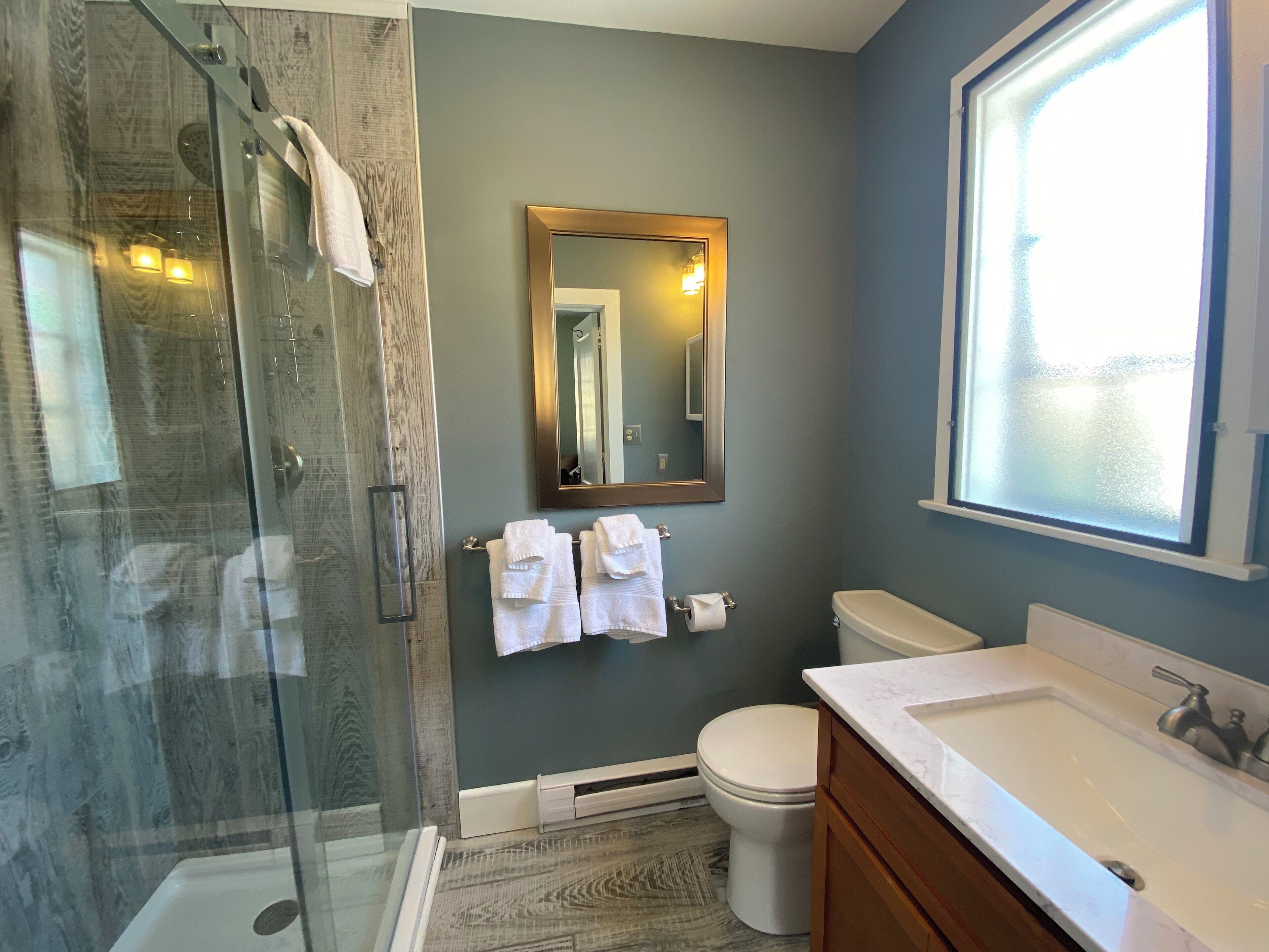 A bathroom with a glass-enclosed shower, a toilet, a sink with a marble countertop, and a mirror with a gold frame.