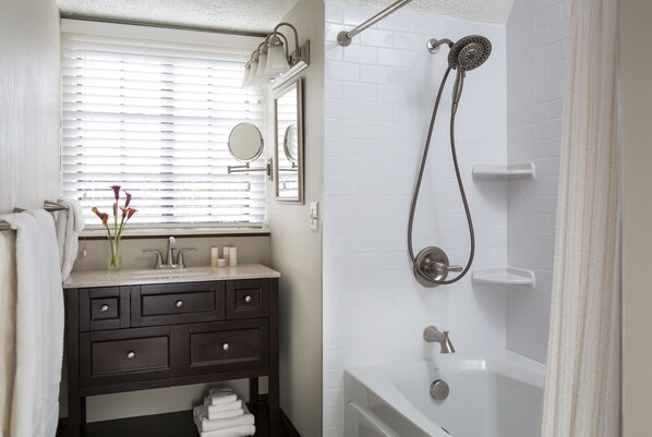 A bathroom with a white bathtub, a dark vanity with drawers, a window with blinds, and a shower with a handheld showerhead. - The Settlers Inn (Hawley)