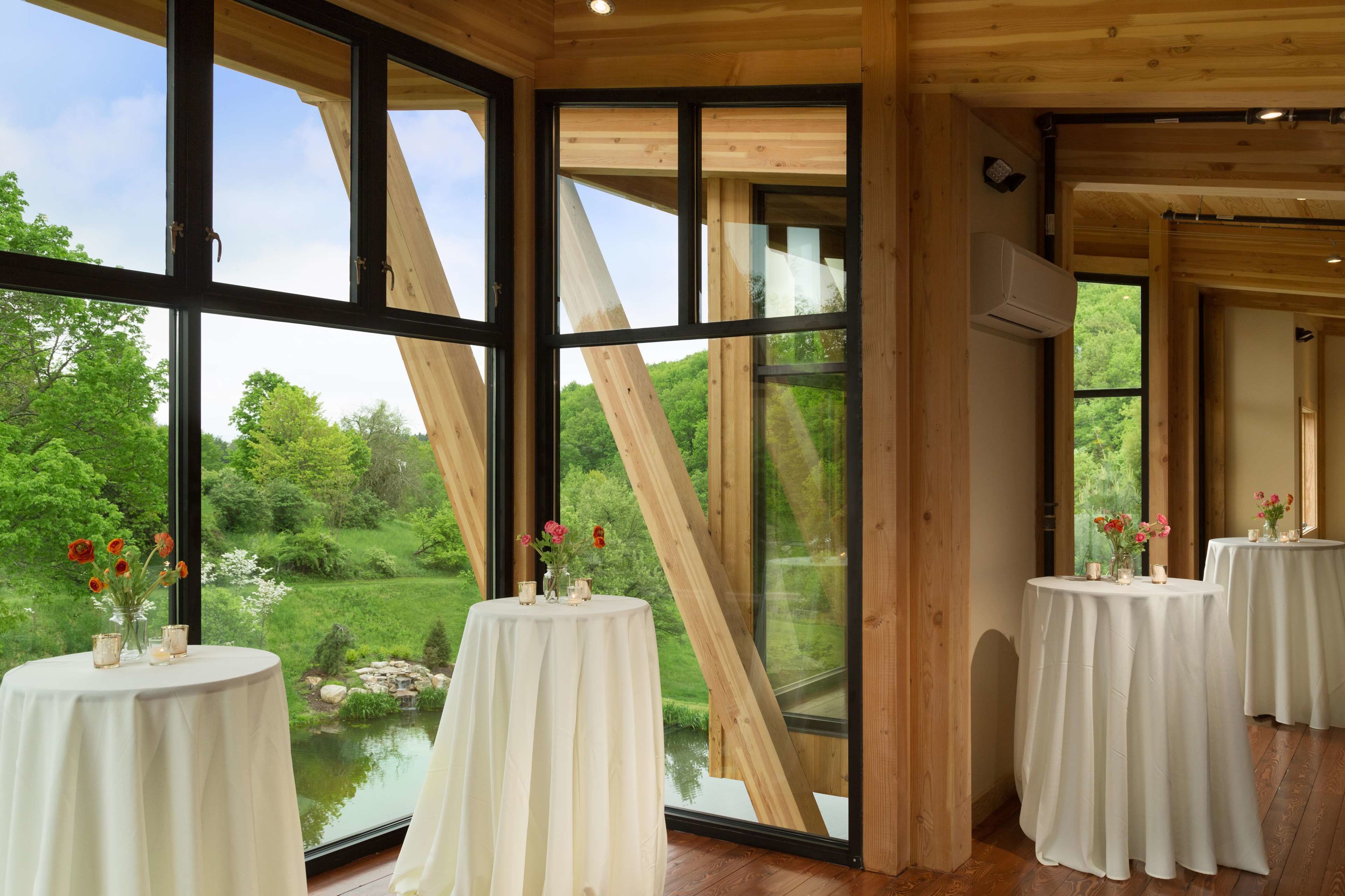 Ein Zimmer mit großen Fenstern und Blick auf einen Teich und Grünflächen. Drei runde Tische mit weißen Tischdecken, jeder Tisch mit einem Blumenstrauß.