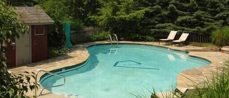 Une piscine dotée d’une plage en pierre, entourée d’arbres et d’un petit abri de jardin.