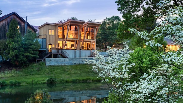 A modern house with a stone foundation, large windows, and a wooden deck overlooking a pond.