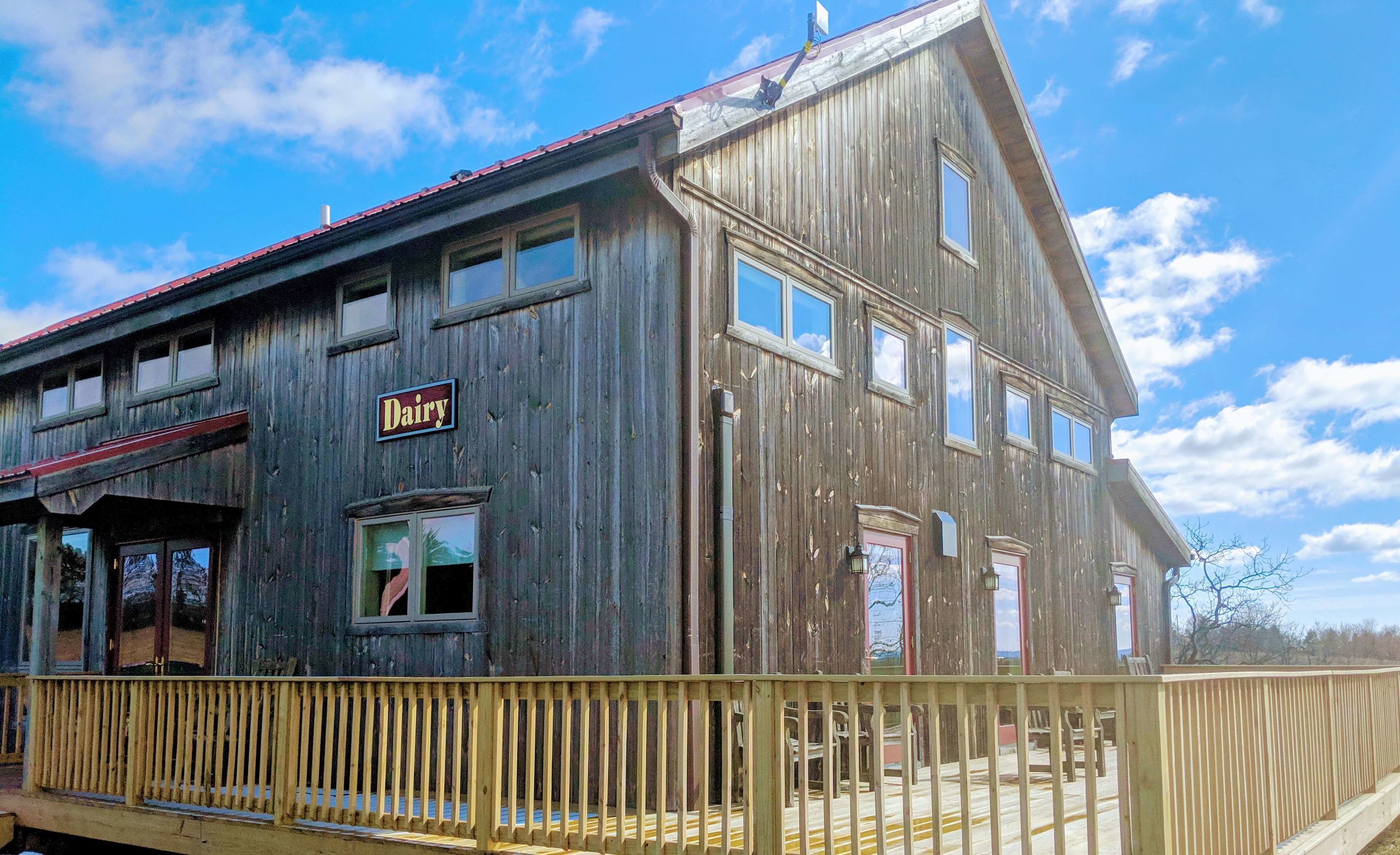 Un bâtiment rustique en bois, avec une enseigne indiquant « Produits laitiers », un porche et une terrasse délimitée par une clôture.