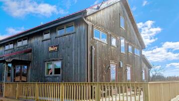 Un bâtiment rustique en bois, avec une enseigne indiquant « Produits laitiers », un porche et une terrasse délimitée par une clôture.