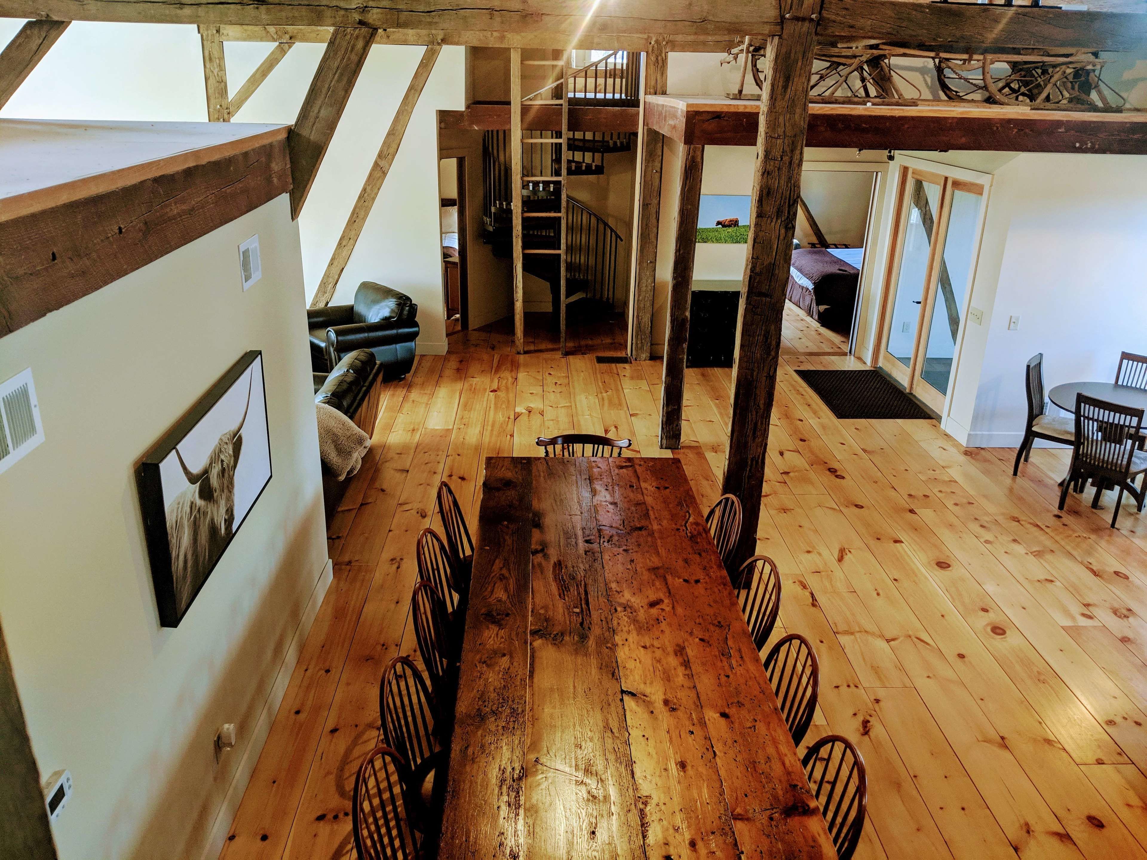 Una habitación amplia con una gran mesa de madera para comer rodeada de sillas, un sofá de cuero negro y una escalera que lleva a un nivel superior.
