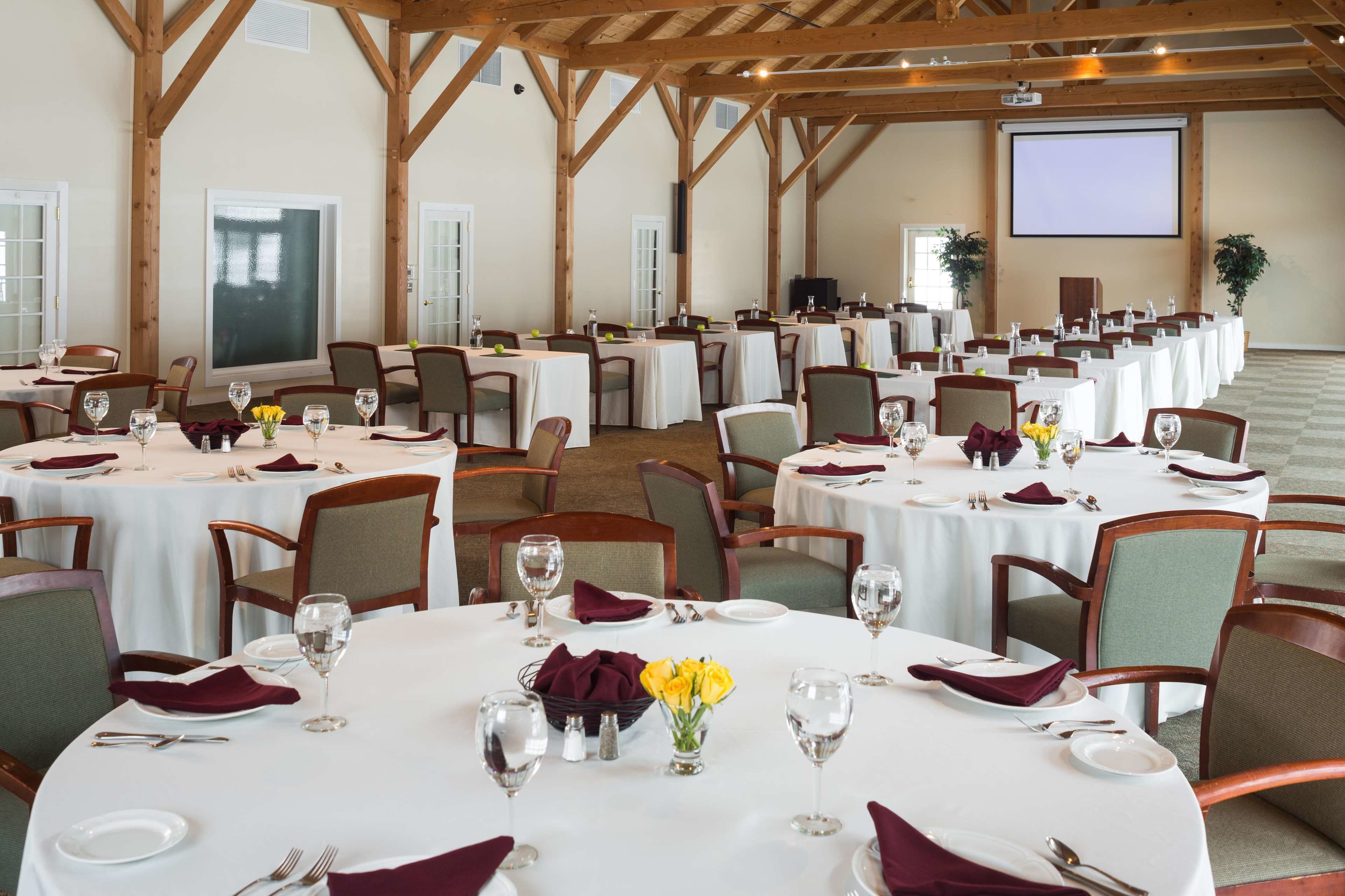 Un salón de banquetes con varias mesas redondas dispuestas para comer, cada una con mantel blanco, servilletas rojas y copas de vino.