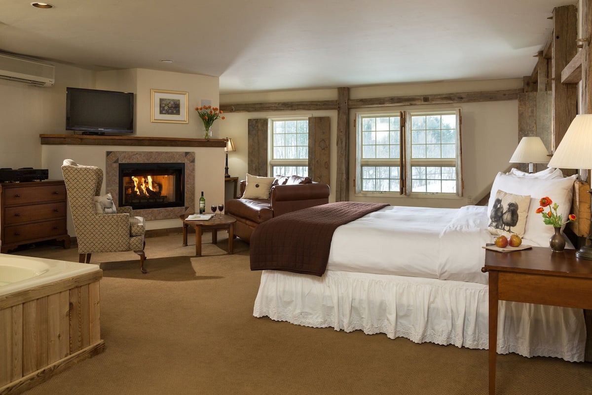 A hotel room with a fireplace, a bed, a sitting area with a chair and a table, a TV, and a window with a view of trees.