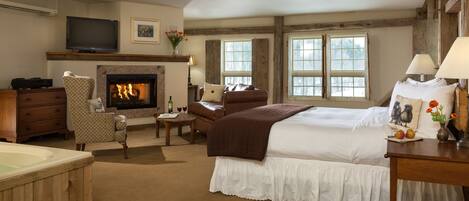 A hotel room with a fireplace, a bed, a sitting area with a chair and a table, a TV, and a window with a view of trees.