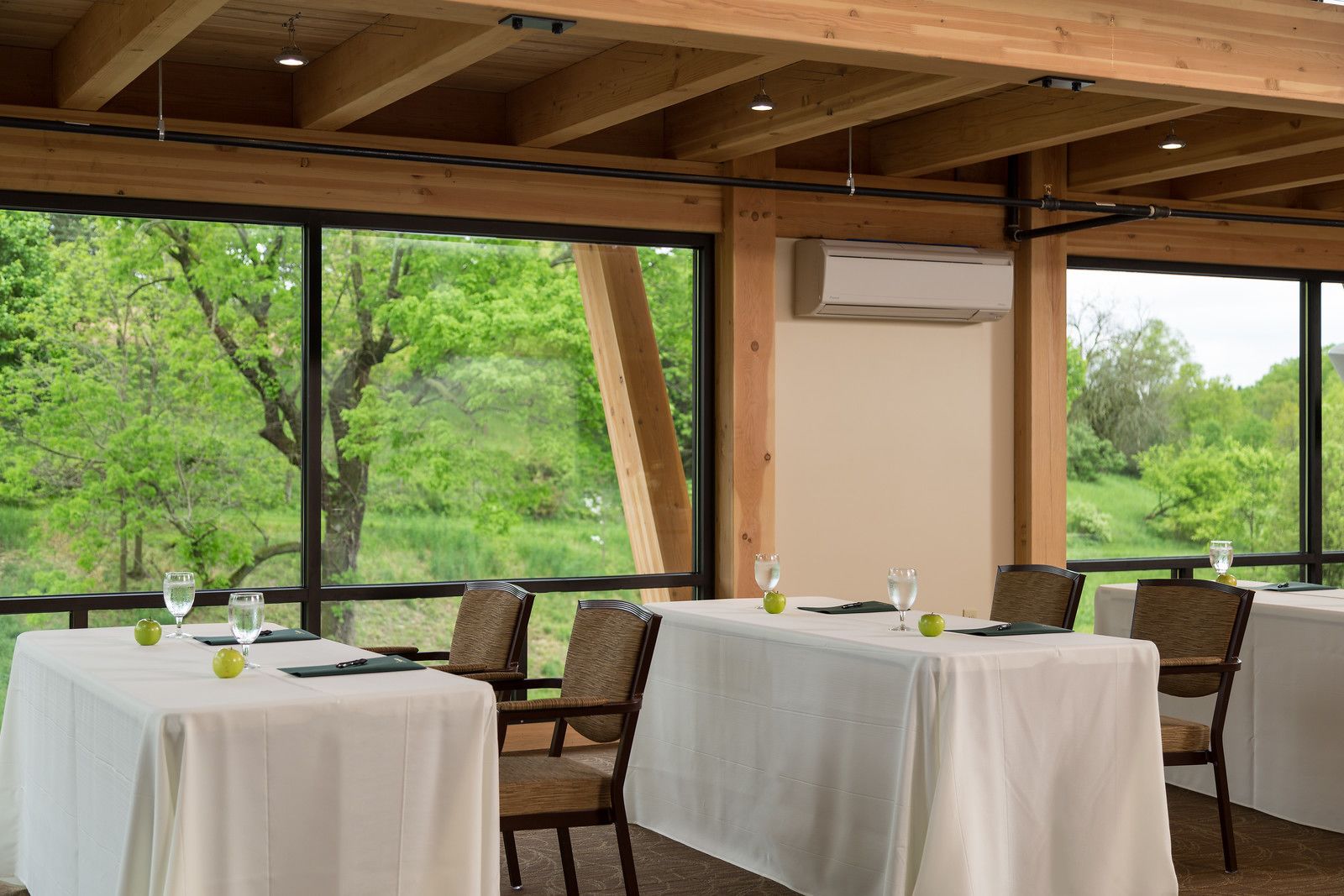A dining area with tables set for two, large windows offering a view of greenery, and an air conditioning unit mounted on the wall.